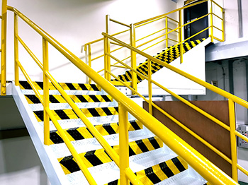 Two sets of stairs lead up to a doorway. The stairs have yellow railing, and the front edge of each tread is marked with yellow and black interlocked chevrons to make them highly visible.